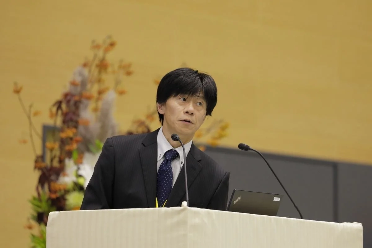 スーツを着た男性が演台でマイクとノートパソコンを前にプレゼンテーションを行っています