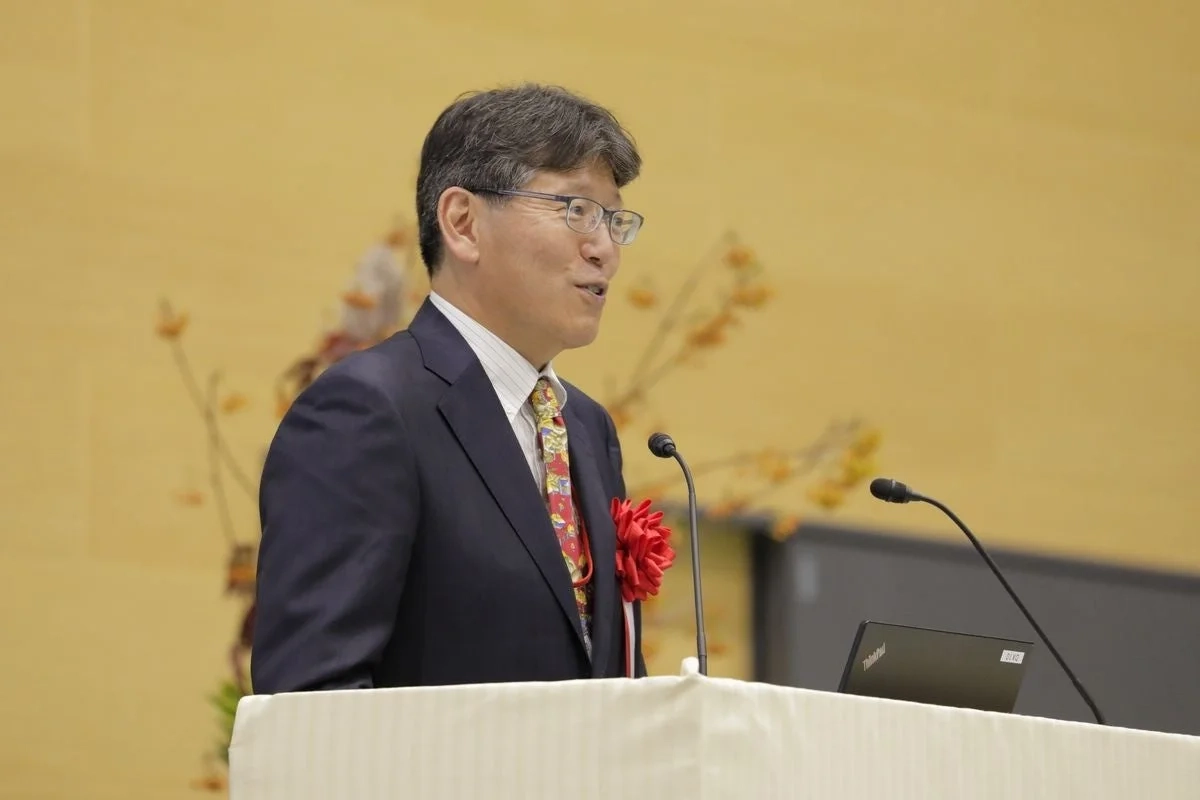 スーツとメガネをかけた男性が、壇上でマイクに向かってスピーチをしている様子