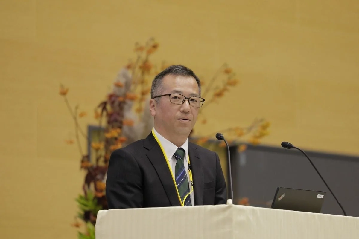 スーツと眼鏡をかけた男性が、壇上でマイクに向かって話している様子