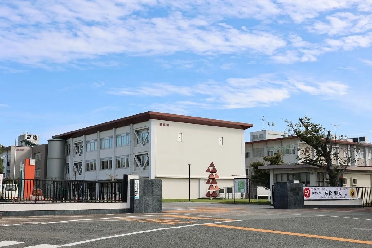 青空の下、高専の校舎と入口が写されており、パリオリンピック出場者である卒業生を祝う横断幕が掲げられています